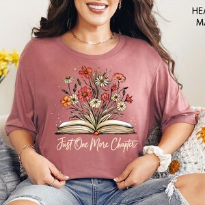 以下が含まれることがあります： ヘザーモーヴのTシャツに、花柄のデザインと「Just One More Chapter」という文字が書かれた開いた本が描かれています。