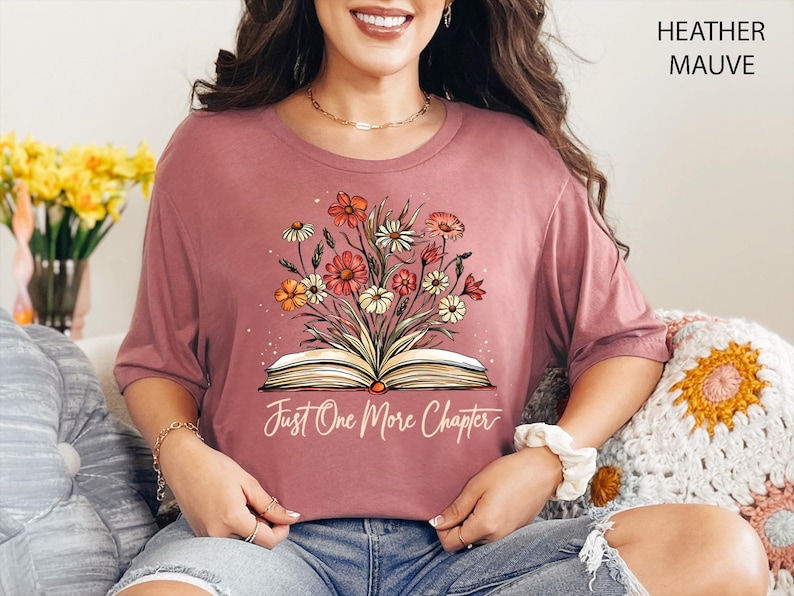 以下が含まれることがあります： ヘザーモーヴのTシャツに、花柄のデザインと「Just One More Chapter」という文字が書かれた開いた本が描かれています。