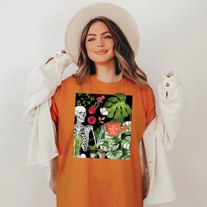 May include: Orange t-shirt with a black and white skeleton graphic surrounded by plants and flowers. The text on the shirt says "You make me feel alive".
