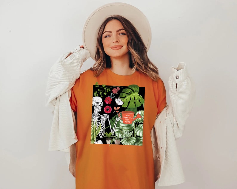 May include: Orange t-shirt with a black and white skeleton graphic surrounded by plants and flowers. The text on the shirt says "You make me feel alive".