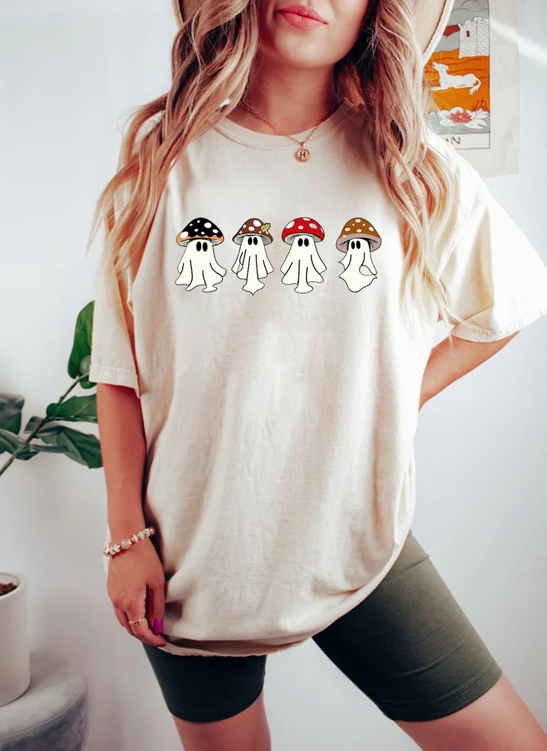 May include: A white t-shirt featuring a design of four ghosts wearing mushroom hats. The mushrooms are in various colours, including red, brown, black, and white.