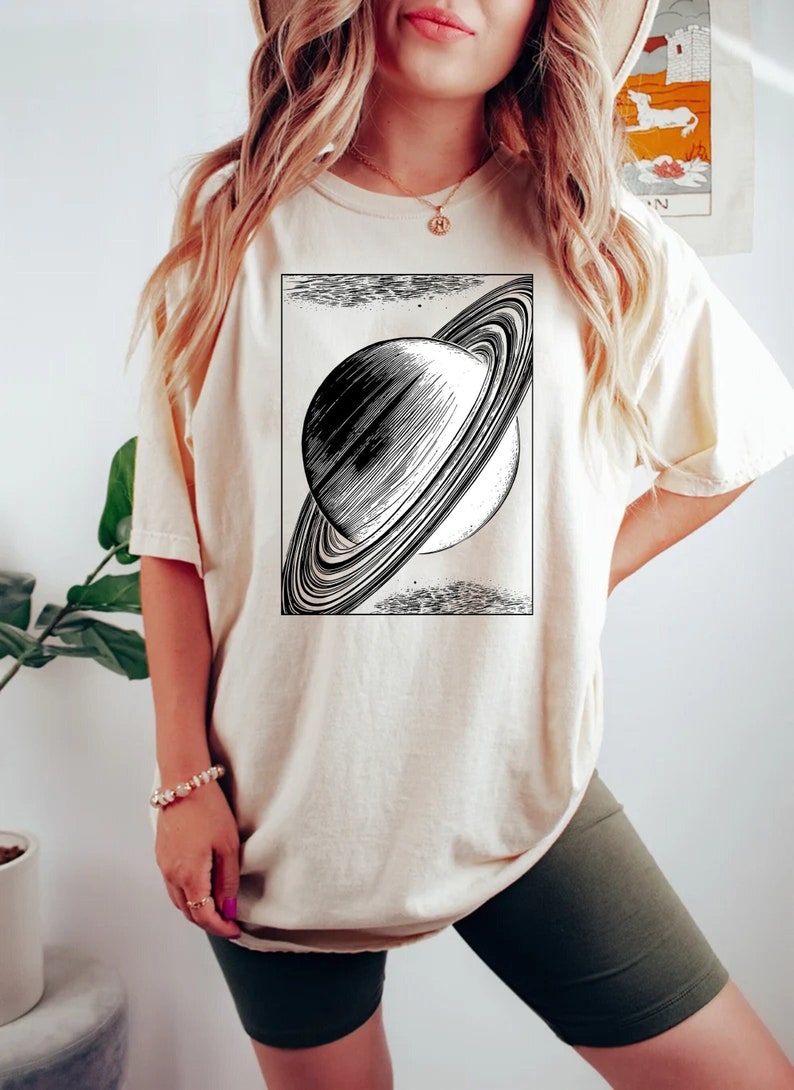 May include: A cream colored t-shirt with a black and white illustration of Saturn with its rings.