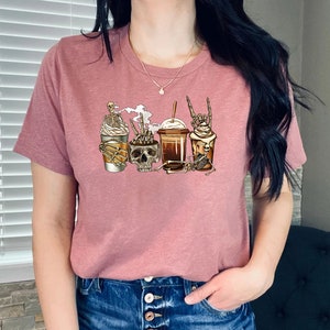 K&ouml;nnte beinhalten: Ein pinkfarbenes T-Shirt mit einem Motiv von vier Kaffeegetr&auml;nken, die mit Skeletten und Sch&auml;deln verziert sind. Die Kaffeegetr&auml;nke umfassen einen Latte, einen Mocha, einen Caramel Macchiato und einen Cold Brew.