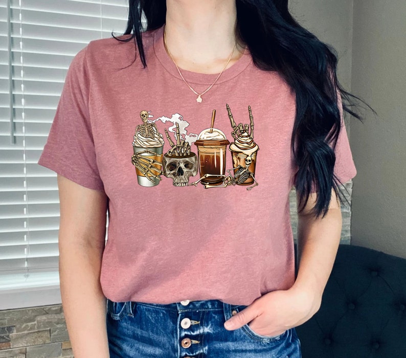 K&ouml;nnte beinhalten: Ein pinkfarbenes T-Shirt mit einem Motiv von vier Kaffeegetr&auml;nken, die mit Skeletten und Sch&auml;deln verziert sind. Die Kaffeegetr&auml;nke umfassen einen Latte, einen Mocha, einen Caramel Macchiato und einen Cold Brew.