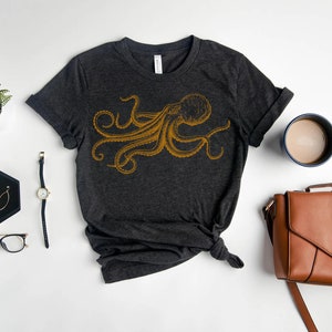 May include: A dark gray t-shirt with a gold octopus graphic. The shirt is laid flat on a white surface with a watch, glasses, earrings, a coffee mug, and a brown leather bag.