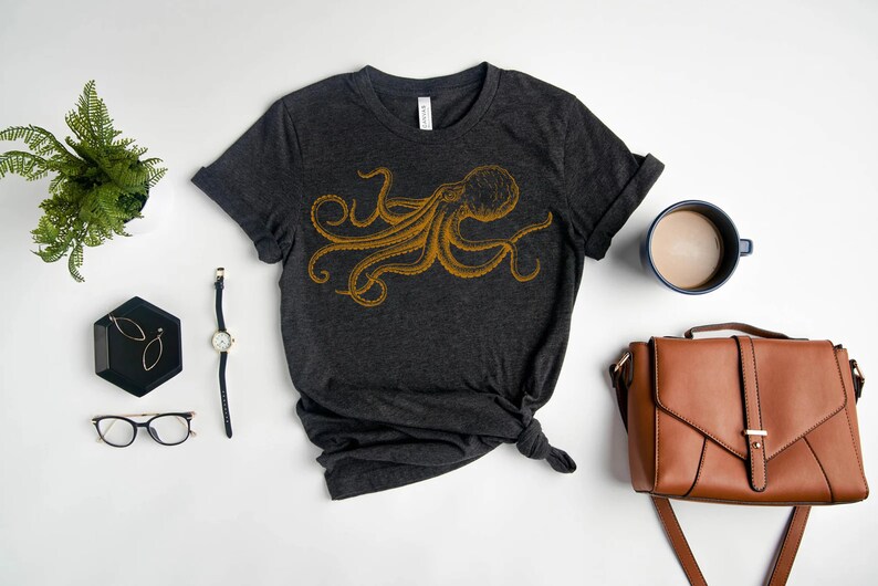 May include: A dark gray t-shirt with a gold octopus graphic. The shirt is laid flat on a white surface with a watch, glasses, earrings, a coffee mug, and a brown leather bag.