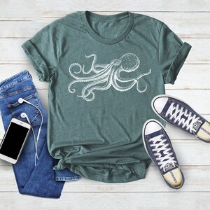 May include: A teal green t-shirt with a white graphic of an octopus. The octopus has eight tentacles and is facing to the right. The t-shirt is laying flat on a white wooden surface.