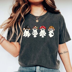 May include: A dark grey t-shirt featuring a graphic of four ghosts wearing mushroom hats. The ghosts are white and the mushrooms are red, brown, and black.