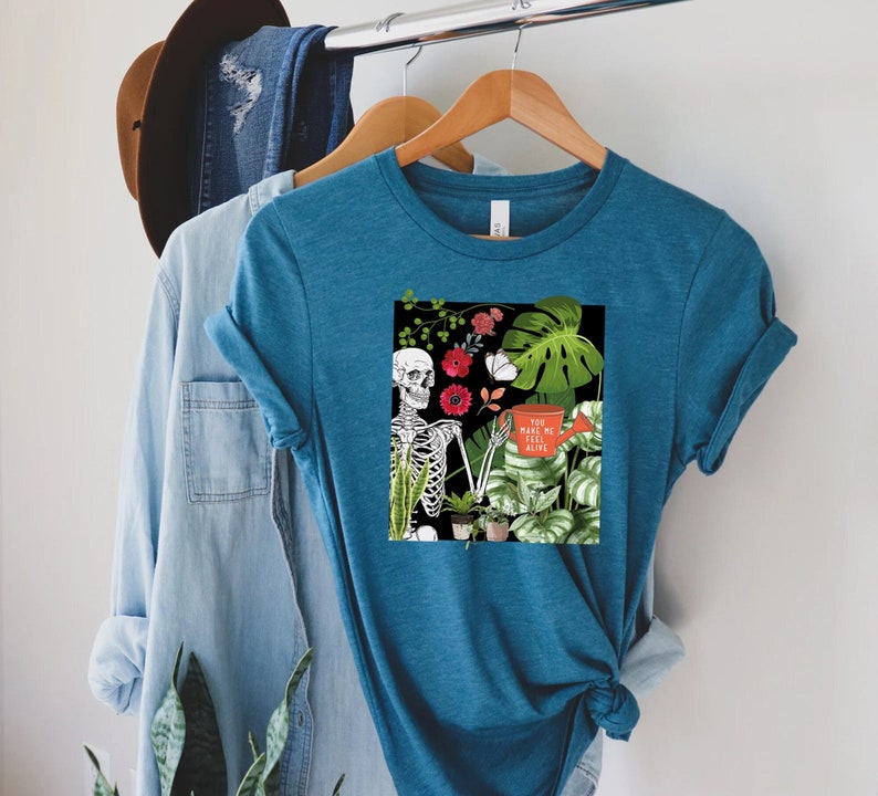 May include: Teal blue t-shirt with a graphic of a skeleton holding a watering can with the text "You make me feel alive". The graphic also includes plants and flowers.
