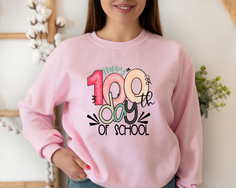 May include: Pink sweatshirt with a graphic design that says "Happy 100th day of school" with colorful donuts forming the number 100.
