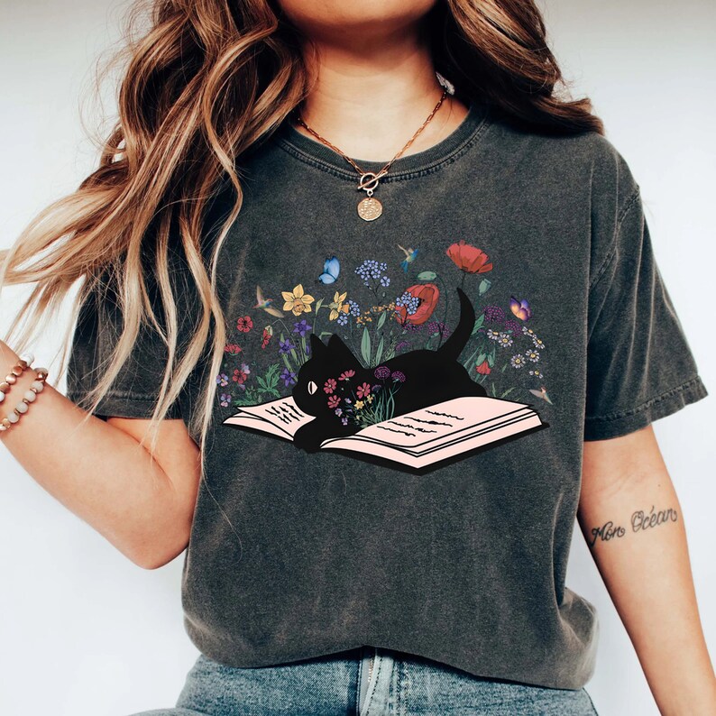 May include: A dark gray t-shirt with a black cat reading a book surrounded by colorful flowers. The cat is lying on an open book with its head resting on the pages. The flowers are a variety of colors, including pink, purple, yellow, and blue.