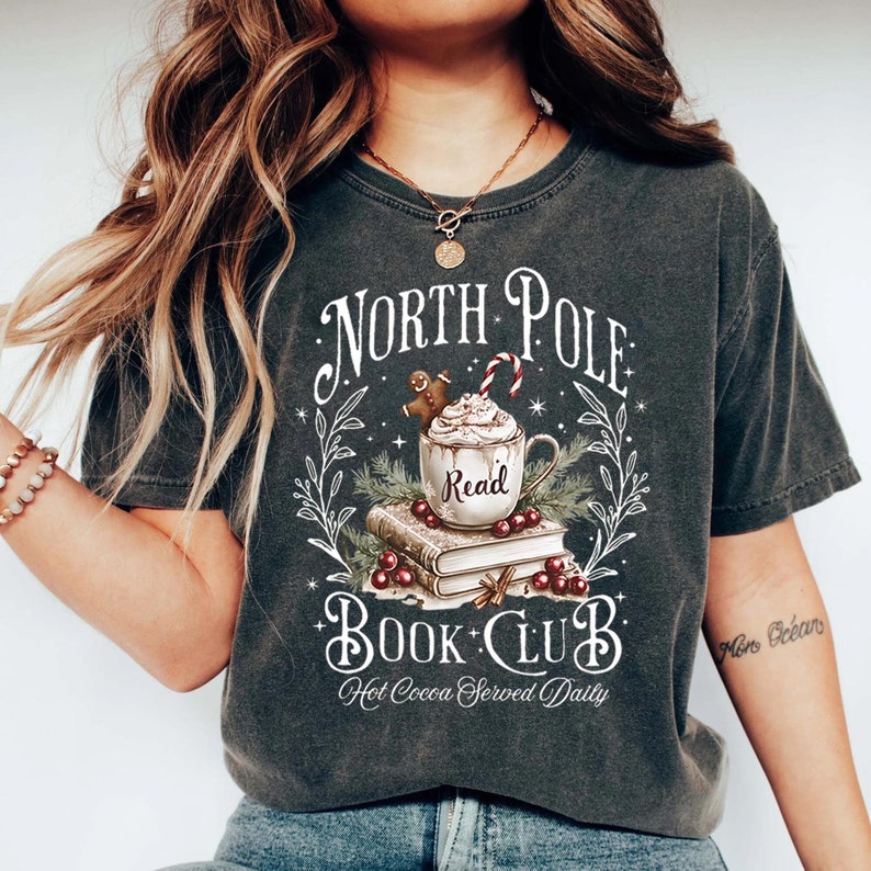 K&ouml;nnte beinhalten: Ein dunkelgraues T-Shirt mit einem wei&szlig;en Grafikdesign, das eine Tasse hei&szlig;er Schokolade mit Schlagsahne und einem Lebkuchenmann zeigt, umgeben von B&uuml;chern und Stechpalmen. Der Text lautet "North Pole Book Club Hot Cocoa Served Daily".