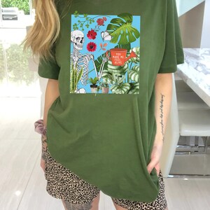May include: A green t-shirt with a graphic of a skeleton holding a watering can and surrounded by plants. The text on the watering can reads "You make me feel alive".