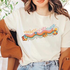 K&ouml;nnte beinhalten: Ein wei&szlig;es T-Shirt mit einem Regenbogen- und Blumendesign und dem Text "Progress is not linear".