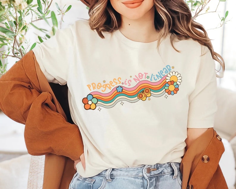 K&ouml;nnte beinhalten: Ein wei&szlig;es T-Shirt mit einem Regenbogen- und Blumendesign und dem Text "Progress is not linear".