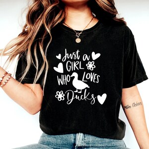 May include: Black t-shirt with the text "Just a Girl Who Loves Ducks" in white. The design includes a duck illustration, hearts, and flower graphics. The shirt is a casual style.
