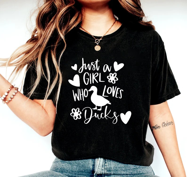 May include: Black t-shirt with the text "Just a Girl Who Loves Ducks" in white. The design includes a duck illustration, hearts, and flower graphics. The shirt is a casual style.