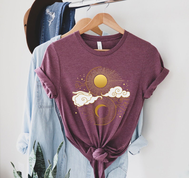 May include: A maroon t-shirt with a gold sun and moon design. The sun and moon are surrounded by clouds and stars.