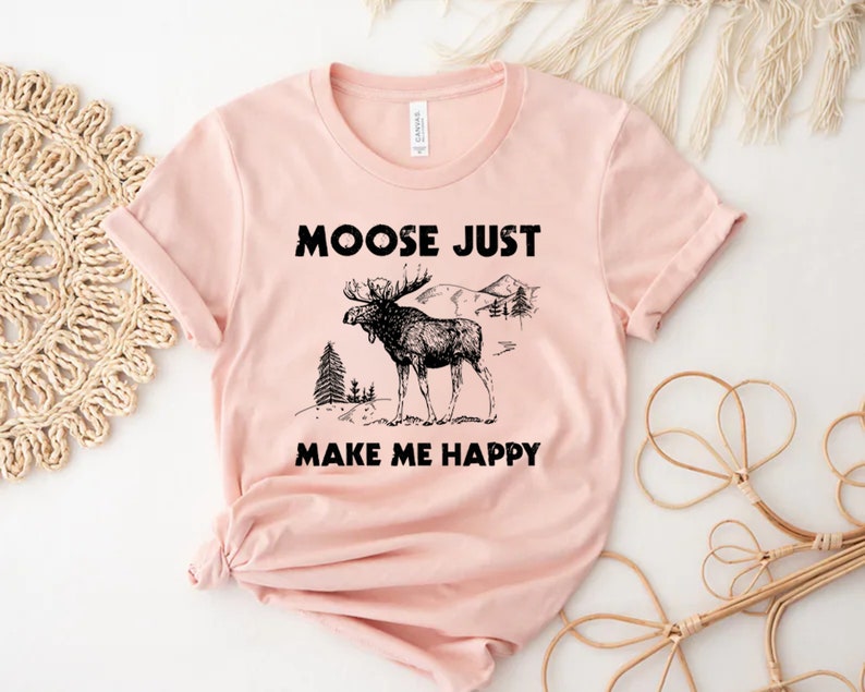 May include: A light pink t-shirt with the text "MOOSE JUST MAKE ME HAPPY" in black. The shirt features a black illustration of a moose with antlers, trees, and mountains.