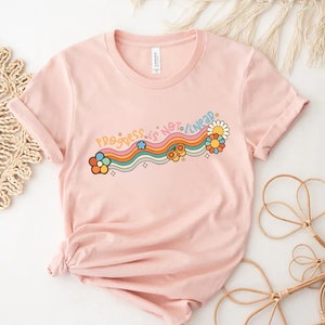 K&ouml;nnte beinhalten: Ein pinkfarbenes T-Shirt mit einem Regenbogen- und Blumendesign. Der Text "Progress is not linear" ist auf das Shirt gedruckt.