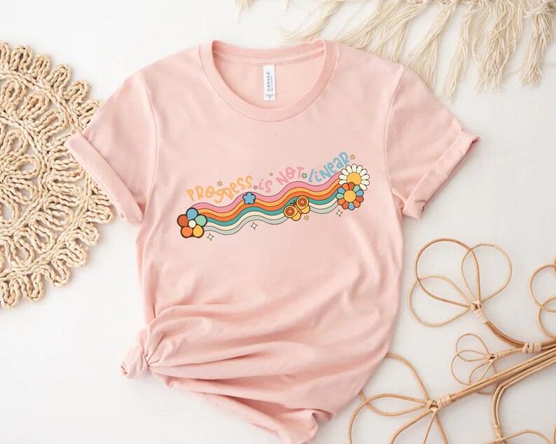 K&ouml;nnte beinhalten: Ein pinkfarbenes T-Shirt mit einem Regenbogen- und Blumendesign. Der Text "Progress is not linear" ist auf das Shirt gedruckt.