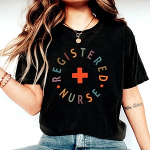 Puede incluir: Una camiseta negra recortada con el texto "REGISTERED NURSE" en un c&iacute;rculo con una cruz roja en el centro. El texto est&aacute; en colores arco&iacute;ris.