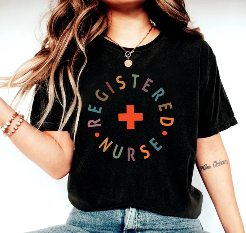 Puede incluir: Una camiseta negra recortada con el texto "REGISTERED NURSE" en un c&iacute;rculo con una cruz roja en el centro. El texto est&aacute; en colores arco&iacute;ris.