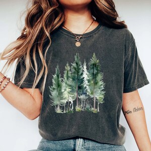Puede incluir: Una camiseta gris oscuro con un estampado de acuarela de una escena de bosque. El estampado presenta varios &aacute;rboles verdes con troncos marrones. La camiseta tiene un corte holgado y cuello redondo.