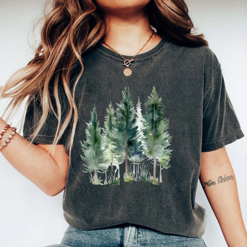 Puede incluir: Una camiseta gris oscuro con un estampado de acuarela de una escena de bosque. El estampado presenta varios &aacute;rboles verdes con troncos marrones. La camiseta tiene un corte holgado y cuello redondo.