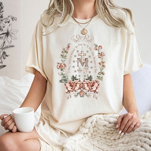 Op de afbeelding: Een wit T-shirt met een bloemenontwerp en de tekst "O Maria, ontvangen zonder zonde, bid voor ons die onze toevlucht tot u nemen" in een cirkel rond een religieus symbool.