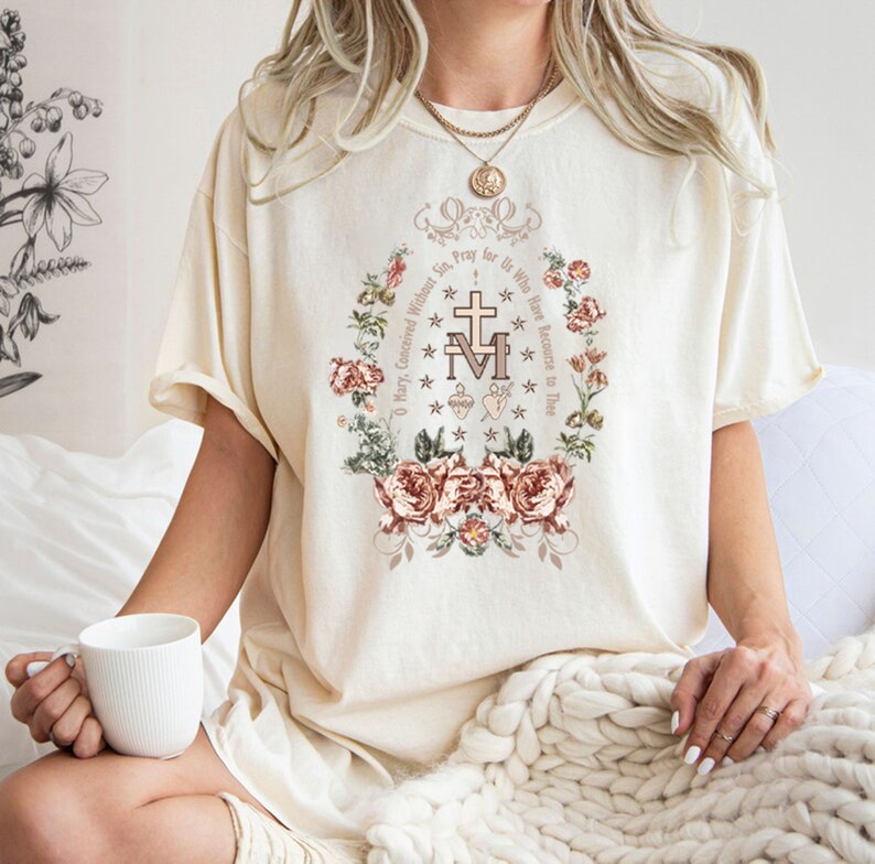 Op de afbeelding: Een wit T-shirt met een bloemenontwerp en de tekst "O Maria, ontvangen zonder zonde, bid voor ons die onze toevlucht tot u nemen" in een cirkel rond een religieus symbool.