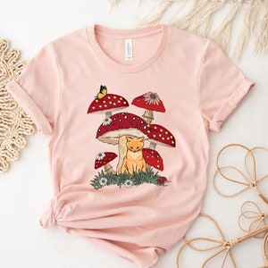 May include: A pink t-shirt with a graphic of a cat sitting under a cluster of red and white mushrooms with a butterfly flying above.