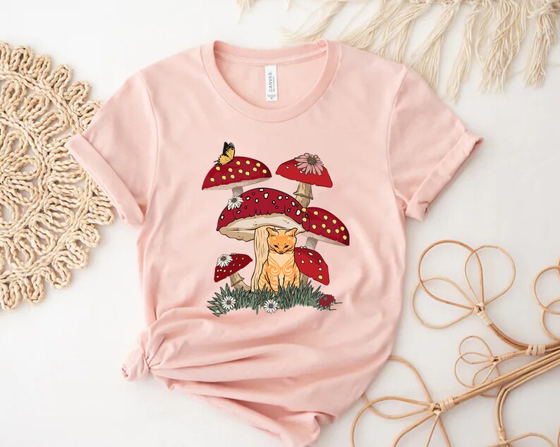 May include: A pink t-shirt with a graphic of a cat sitting under a cluster of red and white mushrooms with a butterfly flying above.