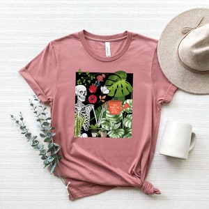 May include: A pink t-shirt with a graphic print of a skeleton holding a watering can with the text "You make me feel alive". The graphic also includes flowers and plants.