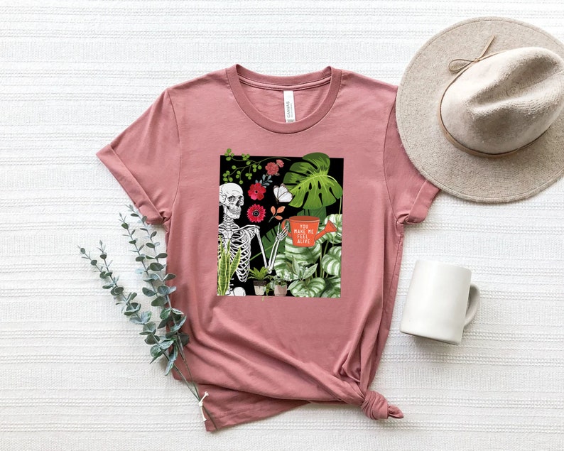 May include: A pink t-shirt with a graphic print of a skeleton holding a watering can with the text "You make me feel alive". The graphic also includes flowers and plants.
