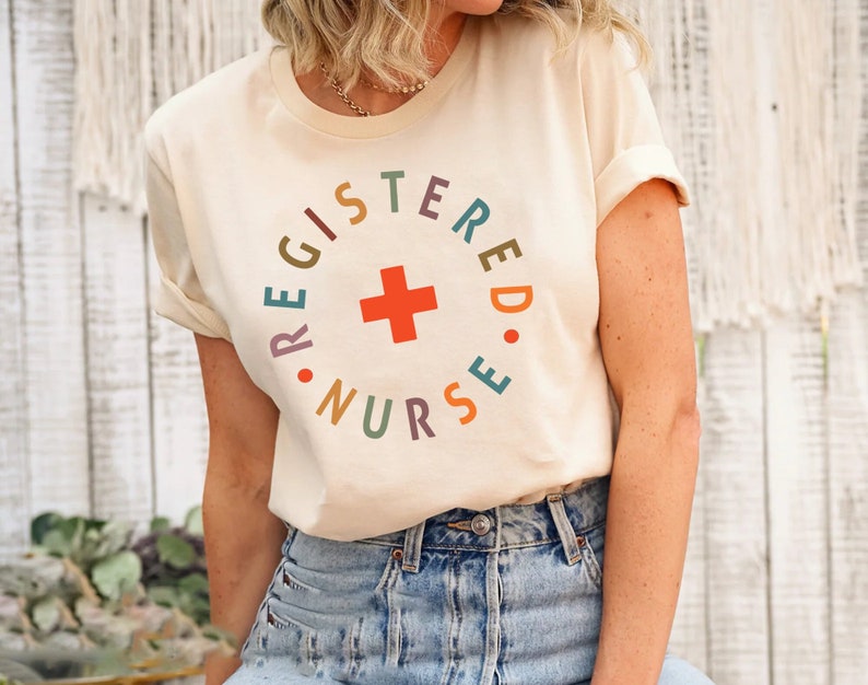 Puede incluir: Una camiseta blanca con un dise&ntilde;o circular de colores que dice "REGISTERED NURSE" con una cruz roja en el centro.