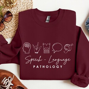 May include: Burgundy sweatshirt with white text and icons that say "Speech-Language Pathology". The icons include a brain, a hand signing, a stack of books, a speech bubble, and an ear.