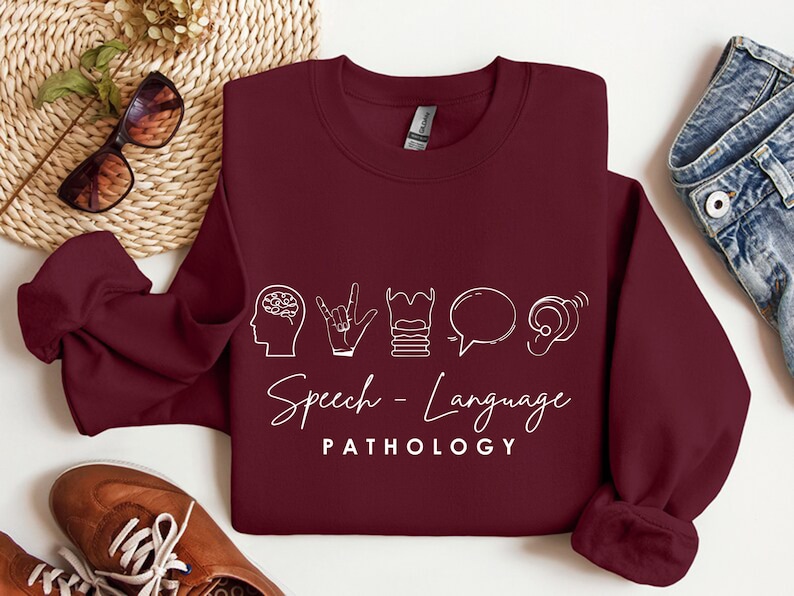 May include: Burgundy sweatshirt with white text and icons that say "Speech-Language Pathology". The icons include a brain, a hand signing, a stack of books, a speech bubble, and an ear.