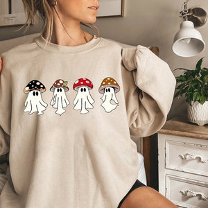 May include: A beige sweatshirt featuring a design of four ghosts wearing mushroom hats. The mushrooms are in various colours and have white spots.