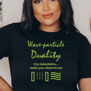 Könnte beinhalten: Ein schwarzes T-Shirt mit grünem Text, der Wave-particle Duality, I'm indecisive... until you observe me lautet. Unter dem Text befinden sich grüne grafische Symbole, die ein Teilchenmuster und ein Wellenmuster auf der Vorderseite darstellen.