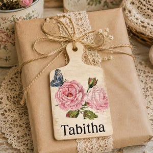 Könnte beinhalten: Ein rechteckiges Geschenkanhänger aus Holz in Form eines kleinen Schneidebretts mit einem Aufdruck von rosa Rosen, grünen Blättern und einem blauen Schmetterling. Der Name Tabitha ist in schwarzer Schrift unten auf dem mit Bindfaden befestigten Anhänger.
