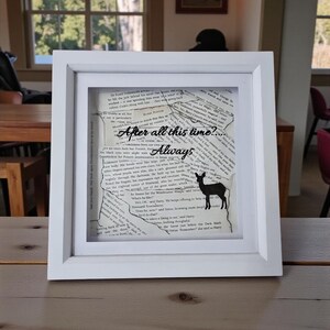 May include: A white framed artwork with torn book pages, the phrases "After all this time? Always", and a black deer silhouette. The piece is displayed on a wooden table, with a window in the background.
