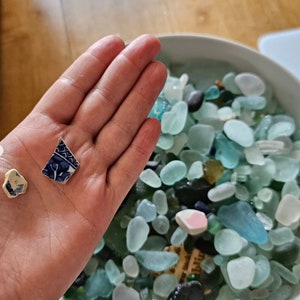 K&ouml;nnte beinhalten: Eine Hand h&auml;lt ein kleines St&uuml;ck blaues und wei&szlig; gemustertes Keramikfliesen. Im Hintergrund befindet sich eine Sch&uuml;ssel gef&uuml;llt mit Seetang in verschiedenen Blaut&ouml;nen und Gr&uuml;nt&ouml;nen.