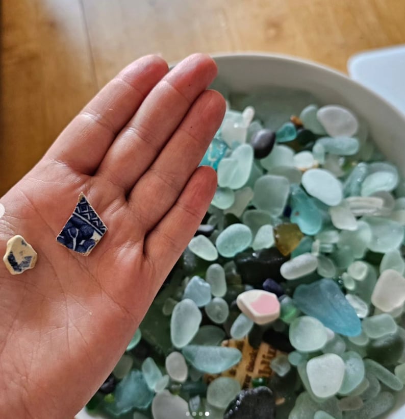 K&ouml;nnte beinhalten: Eine Hand h&auml;lt ein kleines St&uuml;ck blaues und wei&szlig; gemustertes Keramikfliesen. Im Hintergrund befindet sich eine Sch&uuml;ssel gef&uuml;llt mit Seetang in verschiedenen Blaut&ouml;nen und Gr&uuml;nt&ouml;nen.