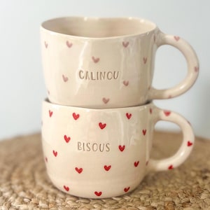Può includere: Due tazze in ceramica impilate, con base color crema e motivi a cuore rossi. La tazza superiore dice "CALINOU" e quella inferiore "BISOUS". Ogni tazza ha un manico arrotondato.