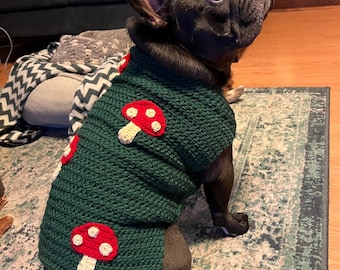 May include: A black French Bulldog wearing a green crochet sweater adorned with red and white mushroom appliqués. The dog is sitting on a patterned rug, with a dark couch and blanket in the background. The sweater is a cozy, handmade item.