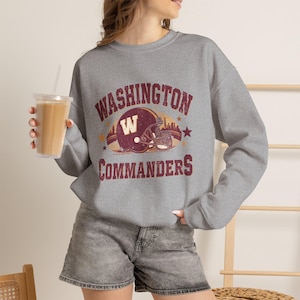 May include: A heather grey crewneck sweatshirt featuring a distressed maroon and gold graphic of a football helmet with the letter W, surrounded by the text WASHINGTON COMMANDERS in a vintage-style font, accented with small stars and a stadium silhouette.