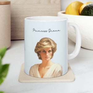 Könnte beinhalten: Weiße Keramiktasse mit hellblauem Hintergrund, die ein Porträt von Prinzessin Diana zeigt. Der Text "Princess Diana." ist in schwarzer Schrift über dem Porträt gedruckt.