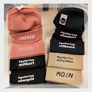 Könnte beinhalten: Eine Auswahl an Mützen in verschiedenen Farben, darunter Rosa, Schwarz und Beige. Jede Mütze hat ein rechteckiges Patch mit Text in Weiß oder Marineblau. Einige Patches haben deutsche Sätze. Die Mützen sind auf einer weißen Oberfläche gestapelt.