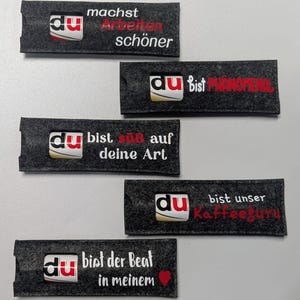 Könnte beinhalten: Fünf dunkelgraue Filztaschen mit dem Wort "du" und verschiedenen deutschen Sätzen. Die Sätze lauten: "machst Arbeiten schöner", "bist phänomenal", "bist süß auf deine Art", "bist unser Kaffeeguru" und "bist der Beat in meinem".
